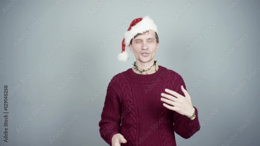 Young man in knitted sweater and santa hat talking about something at camera Christmas preparation Young man in knitted sweater and santa hat talking about something at camera Christmas preparation