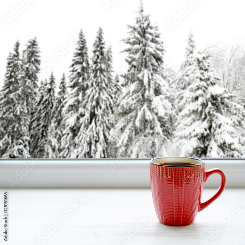 Cup of hot tea on a window sill. In the background, a beautiful winter forest in snow