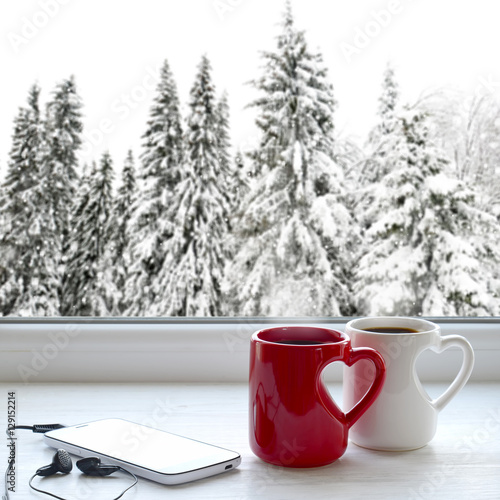 Two cups of coffee, smartphone and headphones on a windowsill. In the background, a beautiful winter forest in snow