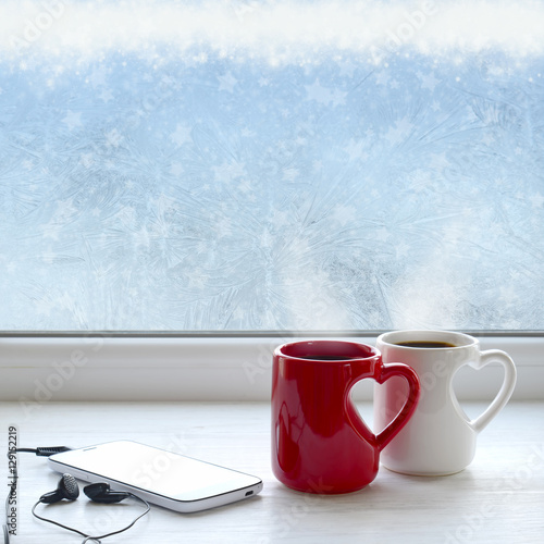 Two cups of coffee  smartphone and headphones on a windowsill. In the background frosty pattern on window as a Christmas background