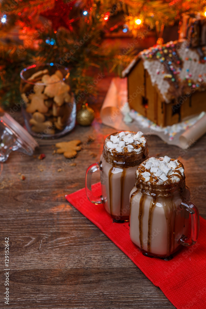 Two glasses of cocoa with marshmallows, whipped cream and chocolate syrup on wooden table and Gingerbread house, cookie jar and christmas tree with toys and garland on background.