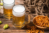 Mugs frothy beer, hops, pretzels and wheat on wooden table