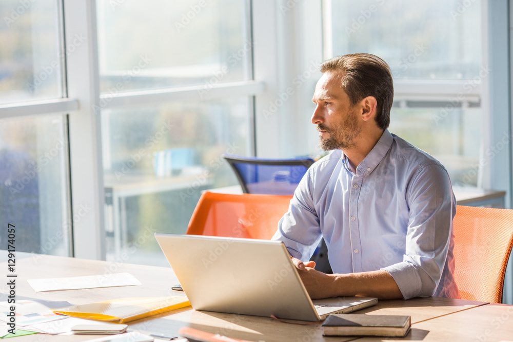 Profile of handsome businessman sitting at table in front of laptop computer and looking at window while working in office interior. Freelance or business concept. Profile of handsome businessman sitting at table in front of laptop computer and looking at window while working in office interior. Freelance or business concept.