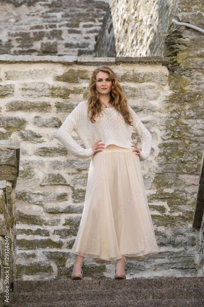 Caucasian white female model and brick stone. Beautiful girl, long red hair, beige skirt and cardigan. Woman standing on the stairs in old town, limestone walls background