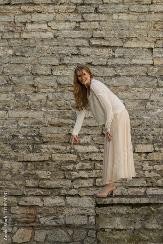 Caucasian white female model and brick stone. Beautiful girl, long red hair, beige skirt and cardigan. Woman standing on the stairs in old town, limestone walls background