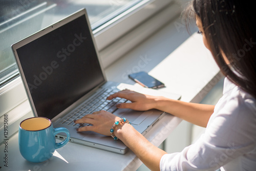 CLoseup picture of businesswoman working on laptop computer with blank screen. Lady drinking tea or coffee while sitting in front of window.