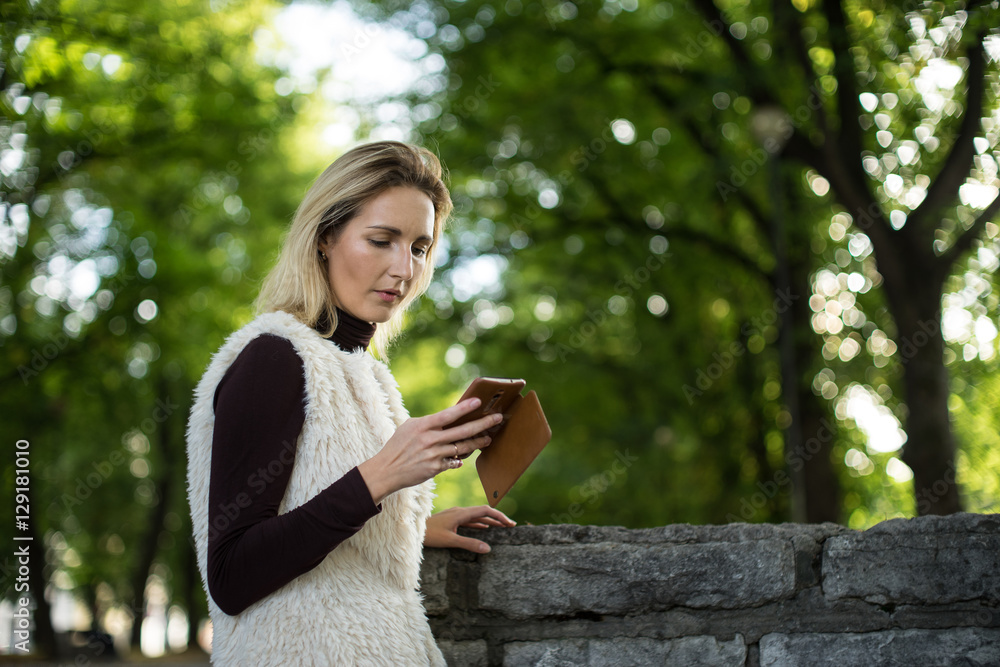Young woman portrait in summer. Blonde girl is reading message on cell phone outside in city nature. Female in a white furry jacket, brown sweater with telephone. Lady dials the number on smartphone. Young woman portrait in summer. Blonde girl is reading message on cell phone outside in city nature. Female in a white furry jacket, brown sweater with telephone. Lady dials the number on smartphone.