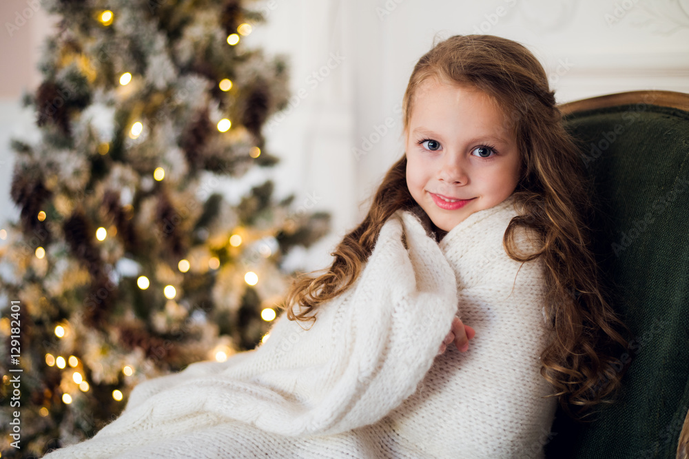 Cute little girl sits by the christmas tree, wrapped in a warm blanket at home Cute little girl sits by the christmas tree, wrapped in a warm blanket at home
