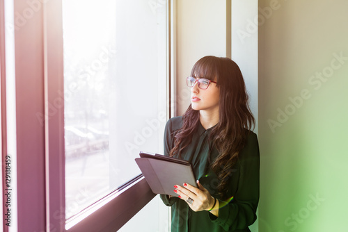 Half length of young handsome caucasian businesswoman near a window holding a tablet, looking down and tapping the screen - business, working, job concept