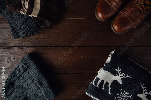 Men's winter clothes on a brown rustic background. Winter shoes, sweater with deer, knitted scarf and hat, gloves. Top view