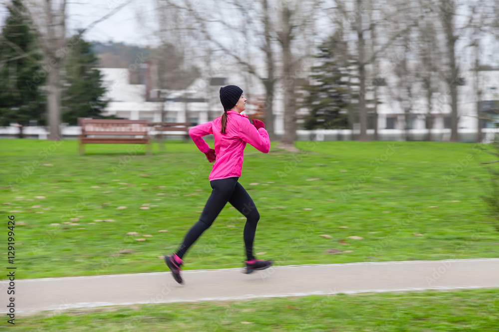 Panning image with motion blur of a young woman running on a cold winter day on an urban park. Profile or side view.