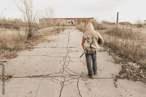 young boy with a gun in his hand. boy goes to an abandoned building. wanderer in the world of the apocalypse. boy in a protective cloak and hood.  a child soldier. young boy soldier. 