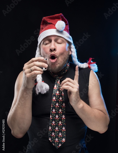 Comic actor man in cap with braids with a glass of vodka on a black background, in anticipation of Christmas and New Year photo