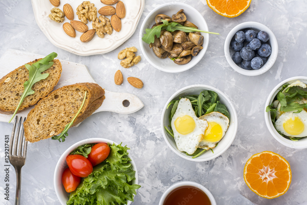 Healthy breakfast with fried eggs, fresh bread, salad, berries, tomatoes, nuts, beverage cup, arugula, mushrooms on a gray light background. Top view Healthy breakfast with fried eggs, fresh bread, salad, berries, tomatoes, nuts, beverage cup, arugula, mushrooms on a gray light background. Top view