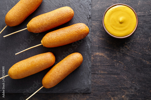 Photography Corn dog traditional American corndog street junk food deep fried hot meat sausage snack with yellow mustard treat coated in a layer of cornmeal batter on stick on black rustic desk