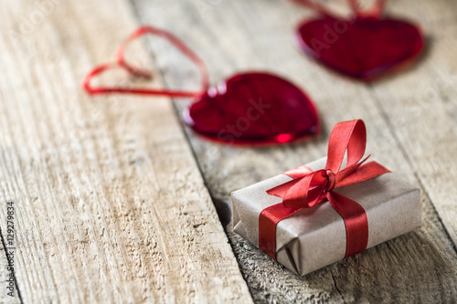 two red glass hearts and little gift wrapped in kraft paper with red shiny silk rape on wooden desk 