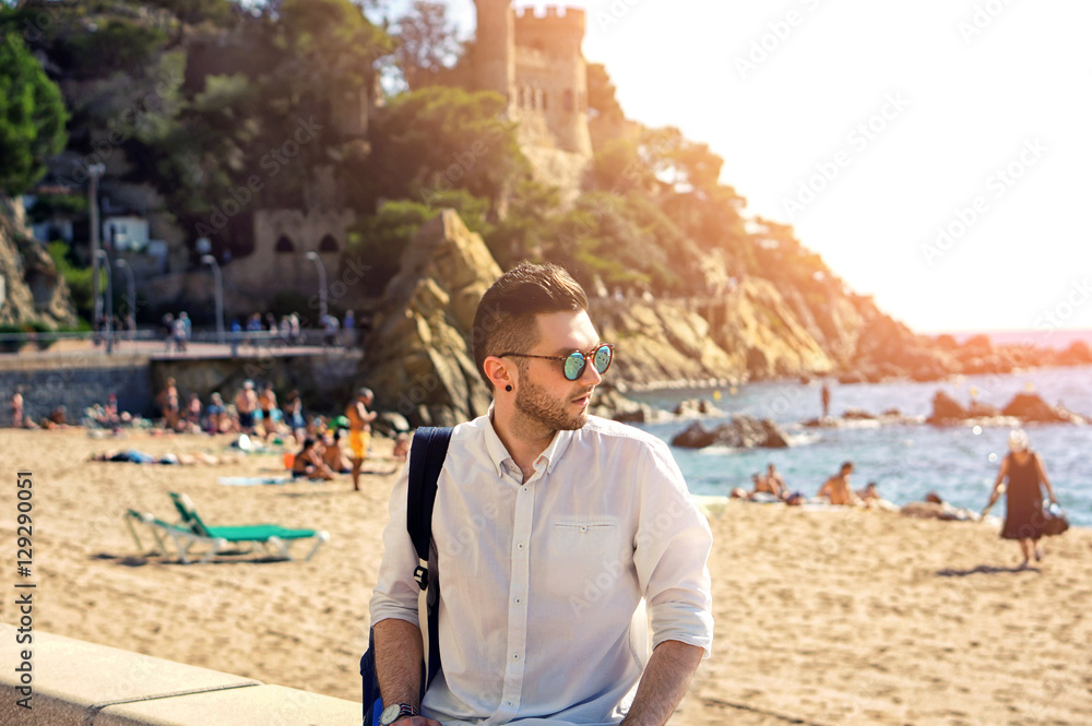 Handsome and confident hipster man in sun glasses with modern haircut and beard, wearing white shirt posing in sea beach scenery in Europe. Beautiful nature and old castle on the background.