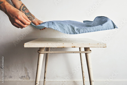 Man with tattooed arms puts a blue and white tablecloth over a small white old table with rust stains