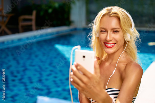 Music and vacation. Enjoying summertime. Pretty young woman in bikini using smartphone in headphones near swimming pool.