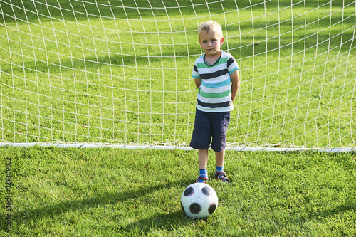 Cute little blond boy playing at being a goalkeeper on a sportsfield standing in the goalposts in sunny summer day