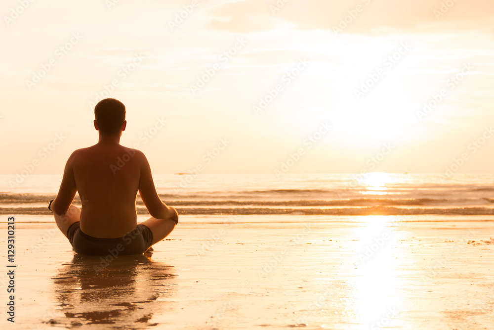 Fototapeta premium Guy is sitting in lotus pose on sunset beach and meditating, enlightenment an zen concept