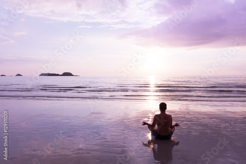 Young man makes meditation in lotus position at sunset on sea beach. Yoga tour or wellness concept