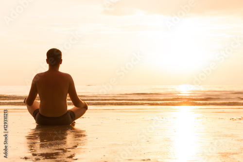 Guy is sitting in lotus pose on sunset beach and meditating, enlightenment an zen concept