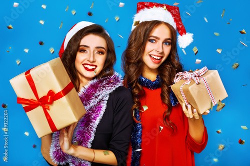 portrait of two happy young women in santa claus hat with gift .Christmas concept. in evening dresses on party over blue background. firecrackers in the background. 