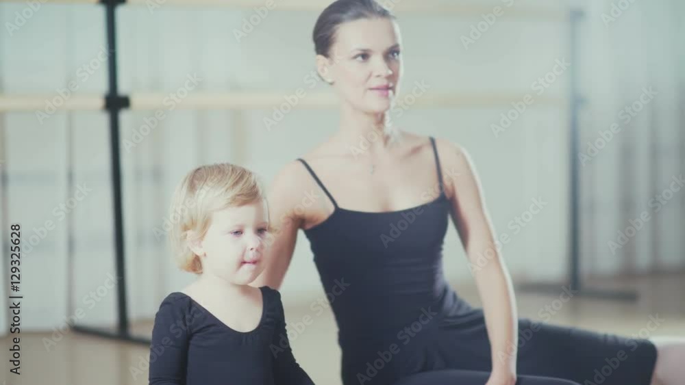 Little Ballerina diligently doing stretching in a ballet class. An adult woman and a little girl ballerina train together. Dance teacher and a little girl sitting on the floor and doing stretching. Little Ballerina diligently doing stretching in a ballet class. An adult woman and a little girl ballerina train together. Dance teacher and a little girl sitting on the floor and doing stretching.