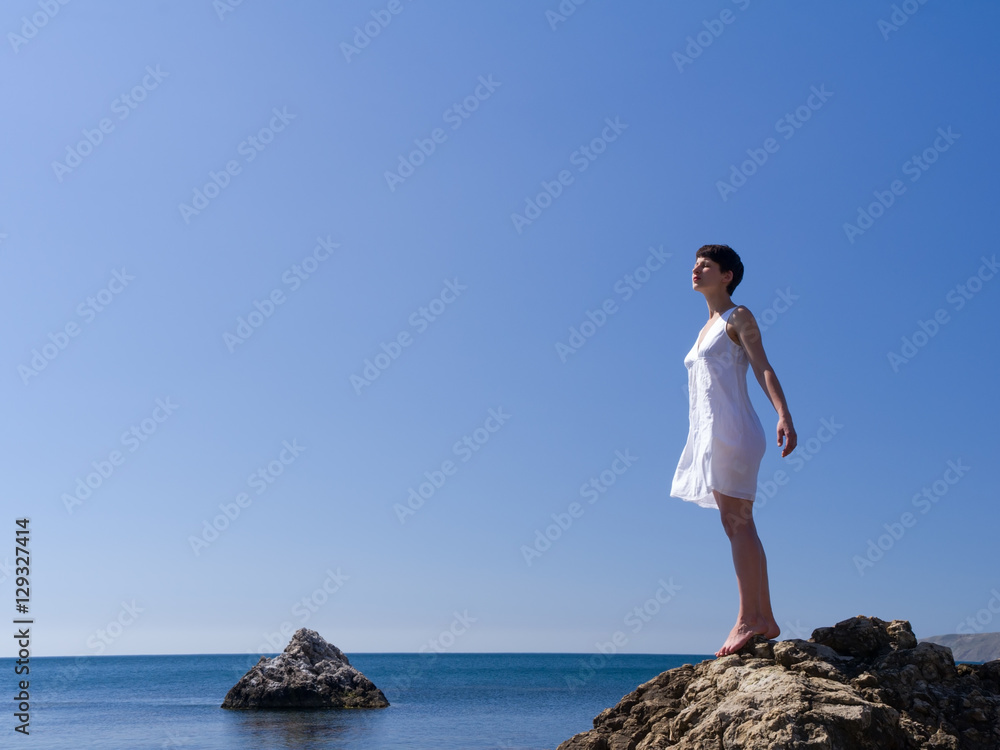 young girl in a white dress stand on the rocks above the sea, her hands like wings