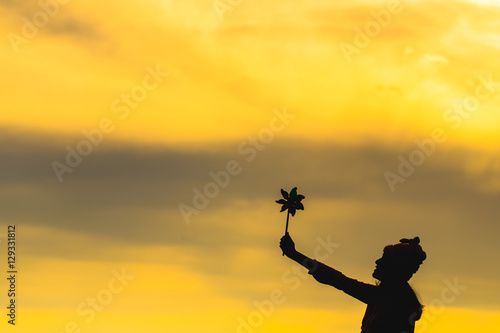 Silhouette of Beautiful girl holding wind toy or wind turbine or pinwheel and wool hat with golden sky on winter season in morning.