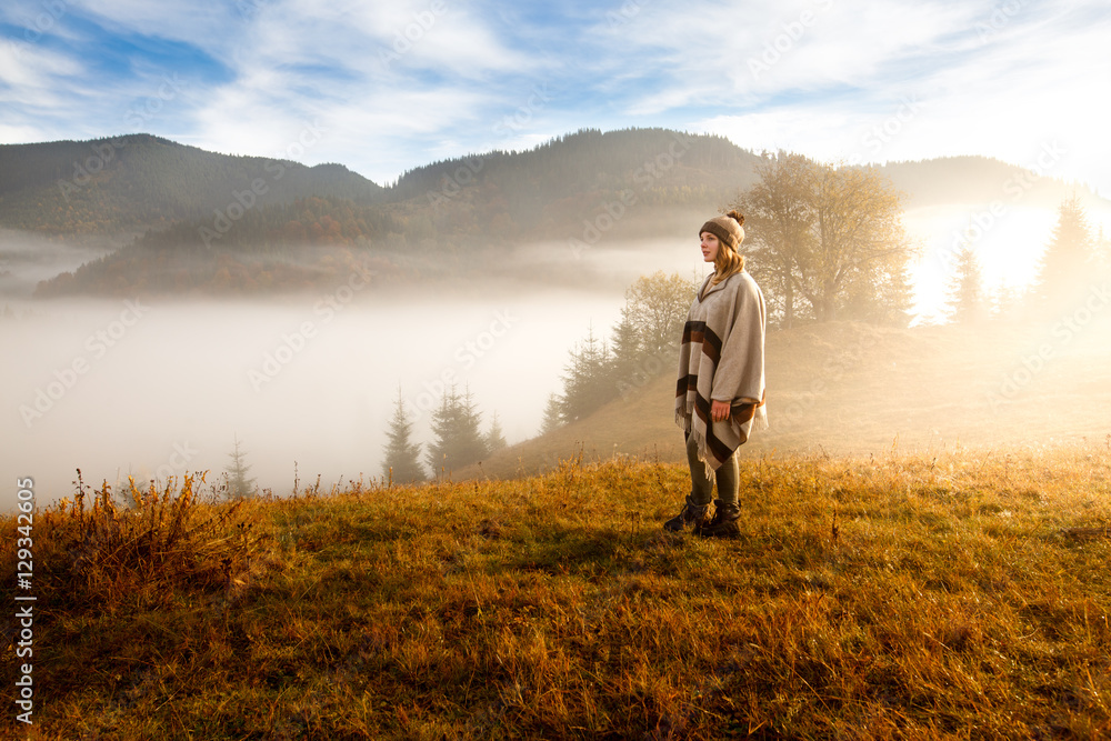 young woman hiking at mountain peak above clouds and fog Hiker girl wrapping in warm poncho outdoor.Early morning.Misty mountain.Young girl over the clouds in the valley looking at calm autumn sunrise