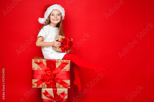 Happy smiling girl in Christmas cap with gift wrapped in craft paper and red ribbons sitting on the gifts on red background. Christmas. Happy family.