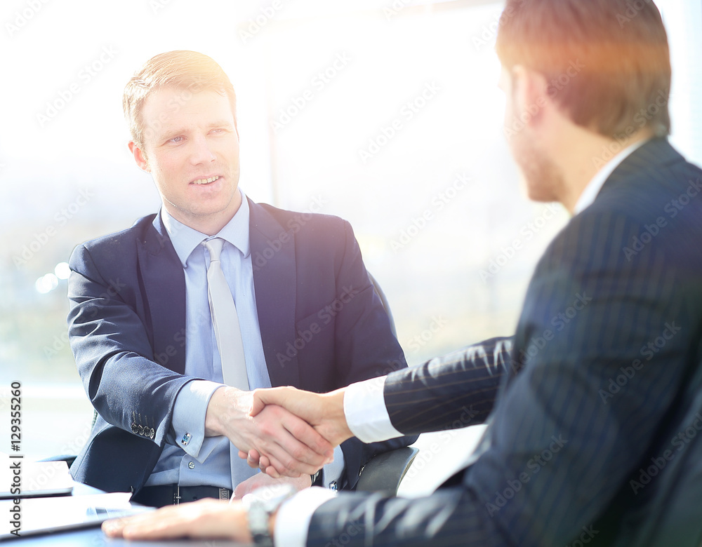 Business colleagues sitting at a table during a meeting with two male executives shaking hands Business colleagues sitting at a table during a meeting with two male executives shaking hands