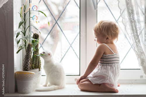 Fototapeta Naklejka Na Ścianę i Meble -  Toddler blonde girl playing with white cat on  window real inter
