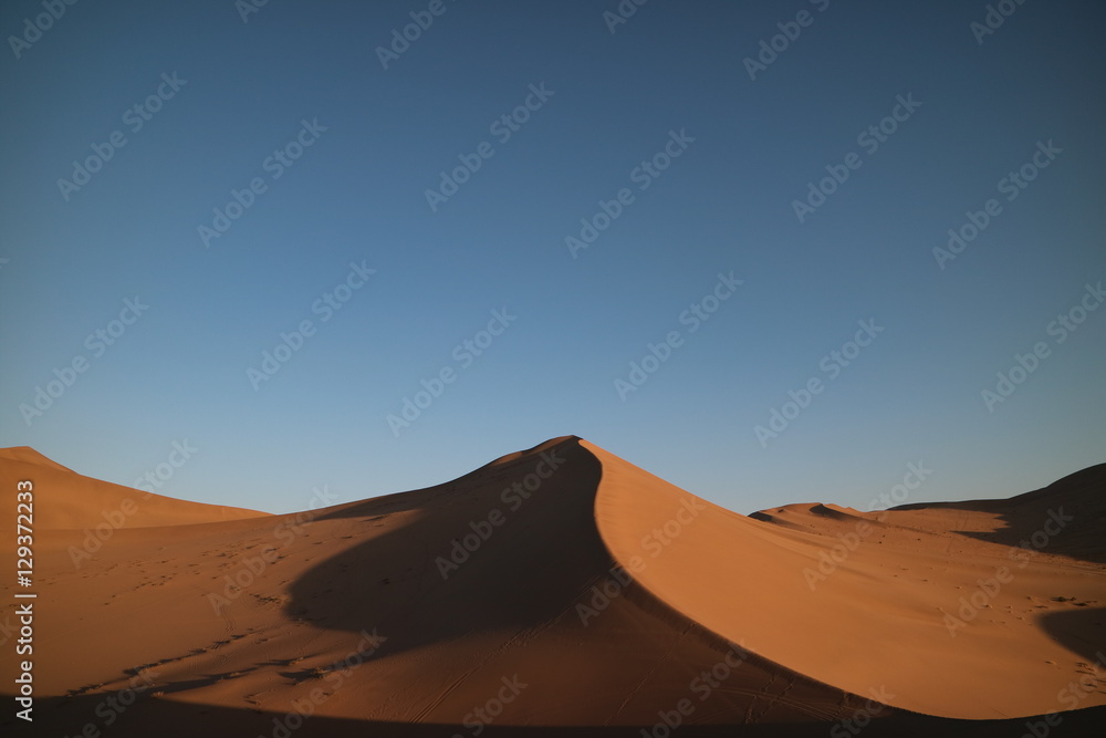 Deserto com grande duna em formato de pico em destaque, sombras projetadas na areia e céu azul