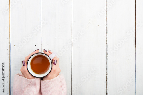 Woman's hands in sweater holding cup of tea on the white wooden table