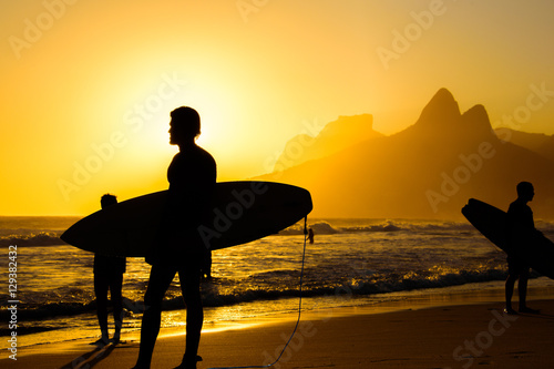 Silhouettes of surfers holding their surfboards on the background of golden sunset on Ipanema Beach and Two Brothers, Dois Irmaos Mountain, Rio de Janeiro, Brazil 