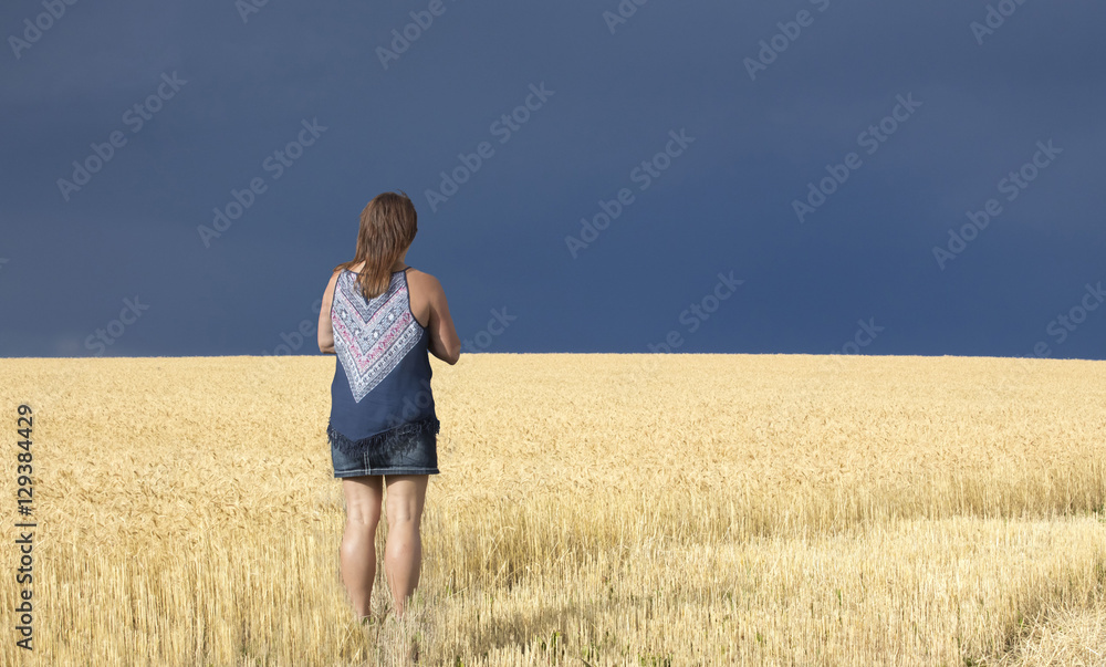 horizontal image of a caucasian woman standing in a field of golden wheat looking toward the sky which is very dark with a thunder storm coming.
