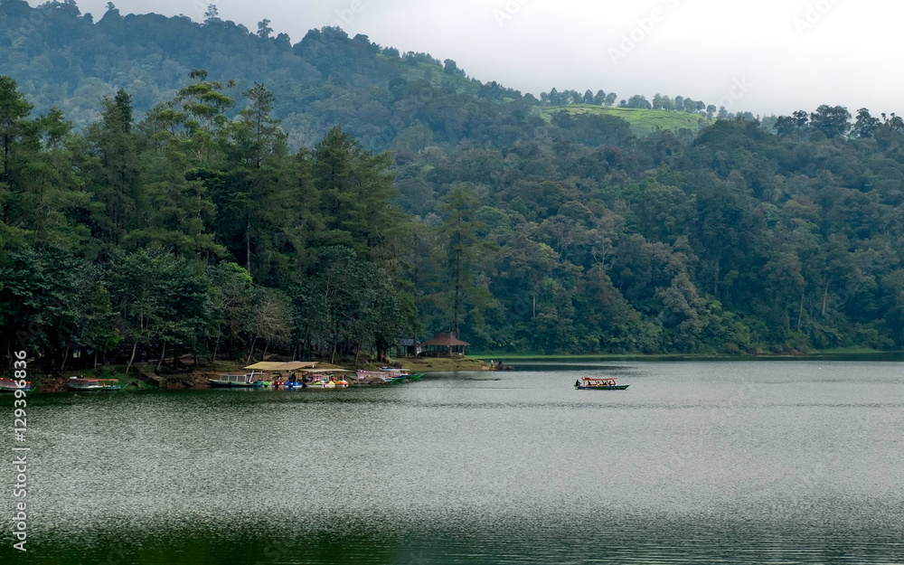 Beautiful scenery of huge lake, with trees, and mist create calming atmosphere. In the middle of the lake, there also a little boat departing from ports, going into the middle of lake