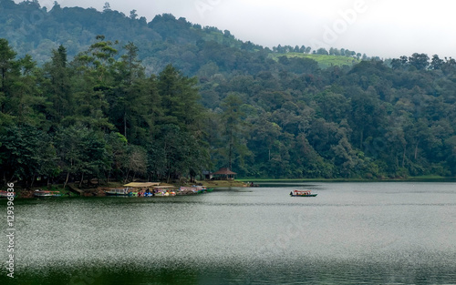 Beautiful scenery of huge lake, with trees, and mist create calming atmosphere. In the middle of the lake, there also a little boat departing from ports, going into the middle of lake