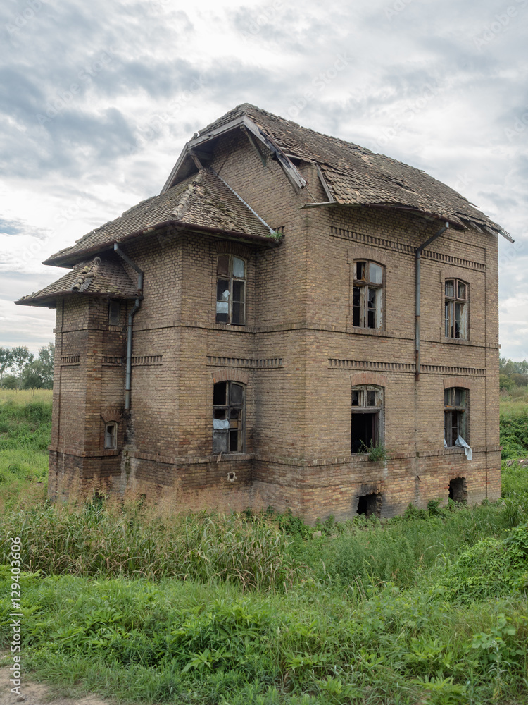 Picture of an old abandoned mansion-house. Verdurous mansion against the background of cloudy sky. Old mansion made of brown bricks. The windows of the mansion are broken.