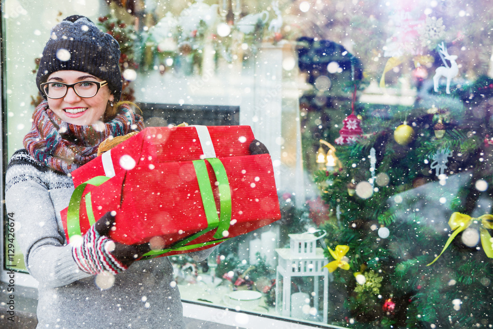 Obraz premium A girl stands at the storefront, on a city street. Shopping, she buys gifts. Woman holding a red box gifts. Background seasonal winter holidays.
