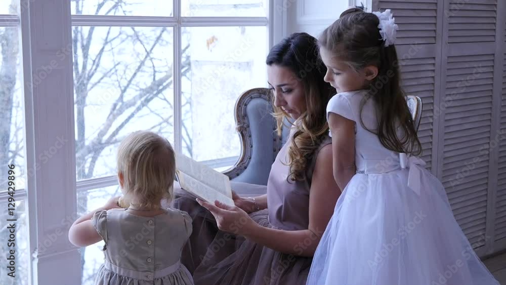 Mother with daughters spending time reading a book. Mom with children reading book near the window Mother with daughters spending time reading a book. Mom with children reading book near the window