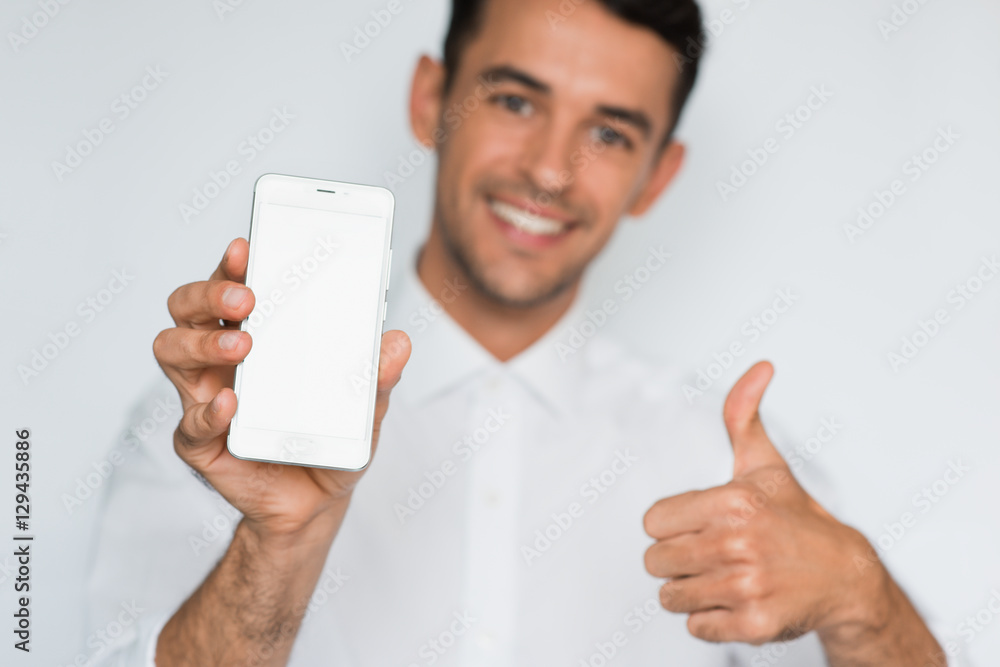 Portrait of young handsome man showing a blank smart phone screen with thumbs up isolated on a light gray background. Smile caucasian businessman with phone in studio. Portrait of young handsome man showing a blank smart phone screen with thumbs up isolated on a light gray background. Smile caucasian businessman with phone in studio.