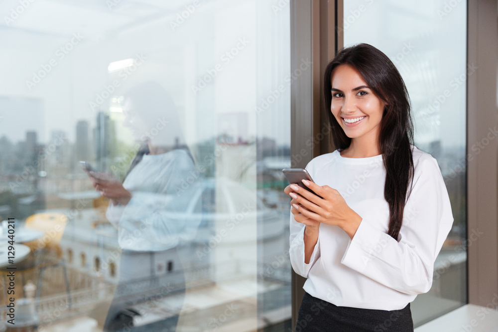 Smiling businesswoman using smartphone near the window in office Smiling businesswoman using smartphone near the window in office