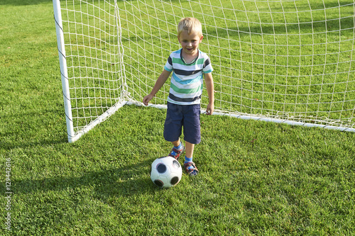 Cute little blond boy playing at being a goalkeeper on a sportsfield standing in the goalposts in sunny summer day