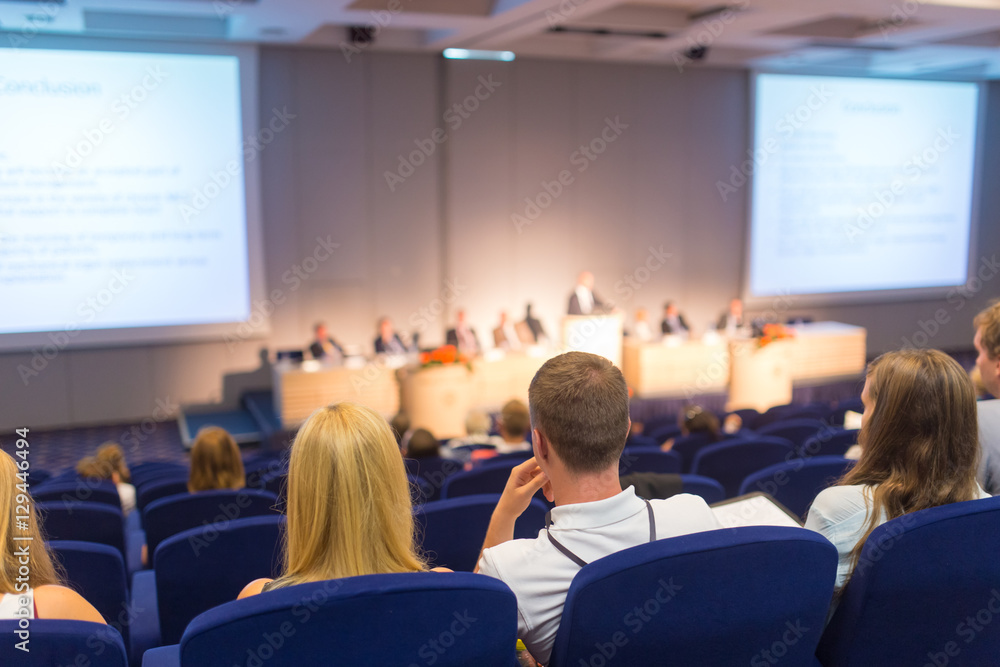 Obraz premium Business and entrepreneurship symposium. Speaker giving a talk at business meeting. Audience in the conference hall. Rear view of unrecognized participant in audience.