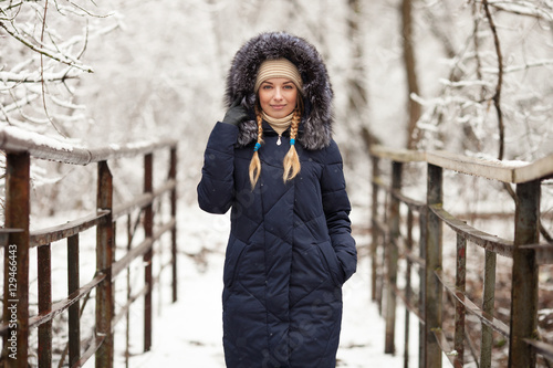 Young adorable woman wearing blue hooded coat enjoying strolling in winter forest outdoors. Nature cold season freshness concept.