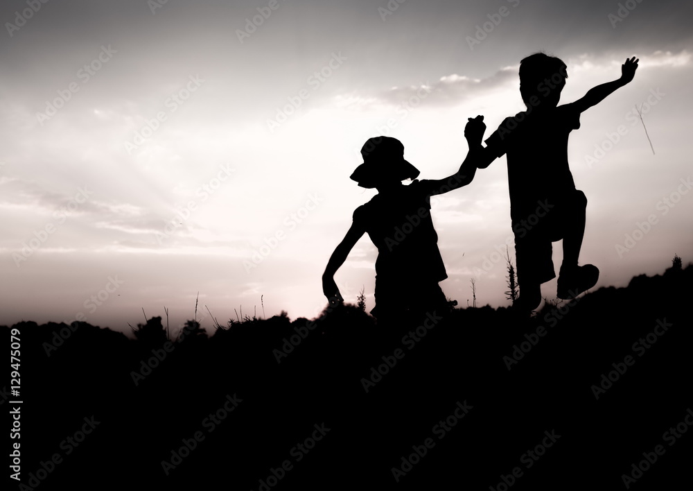 Silhouettes of kids jumping off a cliff at sunset. Boy and girl jump high holding hands. Brother and sister having fun in summer. Friendship, freedom concept. Vacation in mountains. Silhouettes of kids jumping off a cliff at sunset. Boy and girl jump high holding hands. Brother and sister having fun in summer. Friendship, freedom concept. Vacation in mountains.